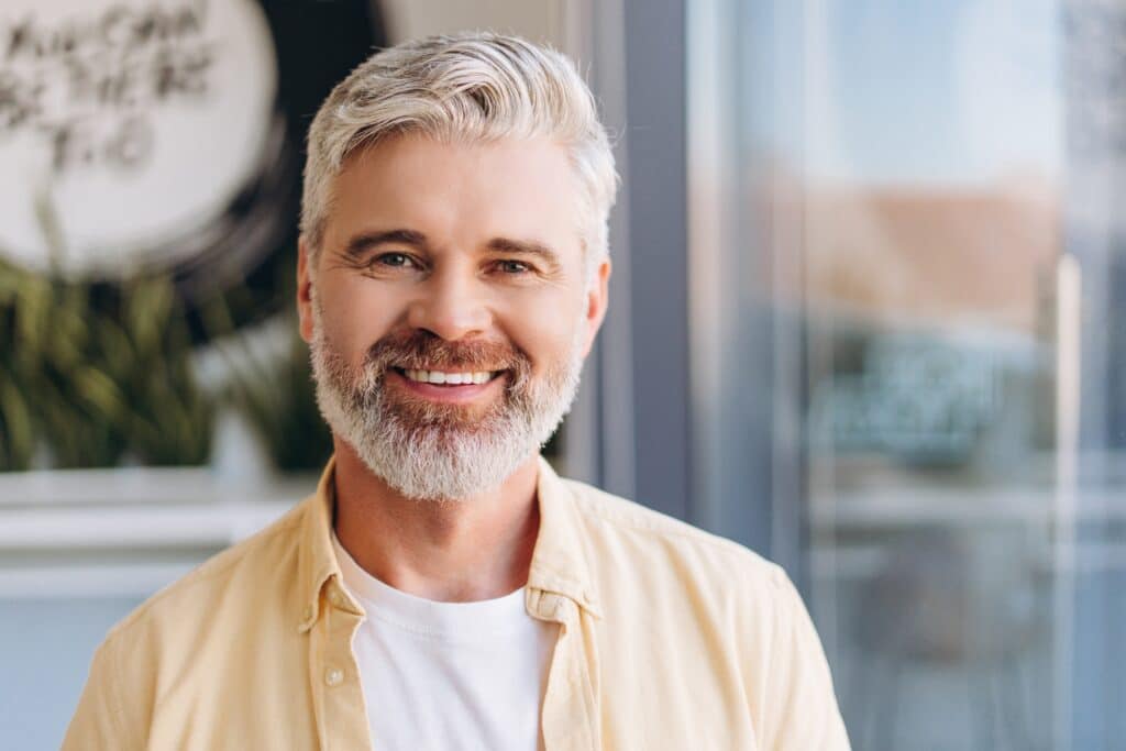 Smiling man with gray hair and beard wearing a yellow shirt, standing indoors.